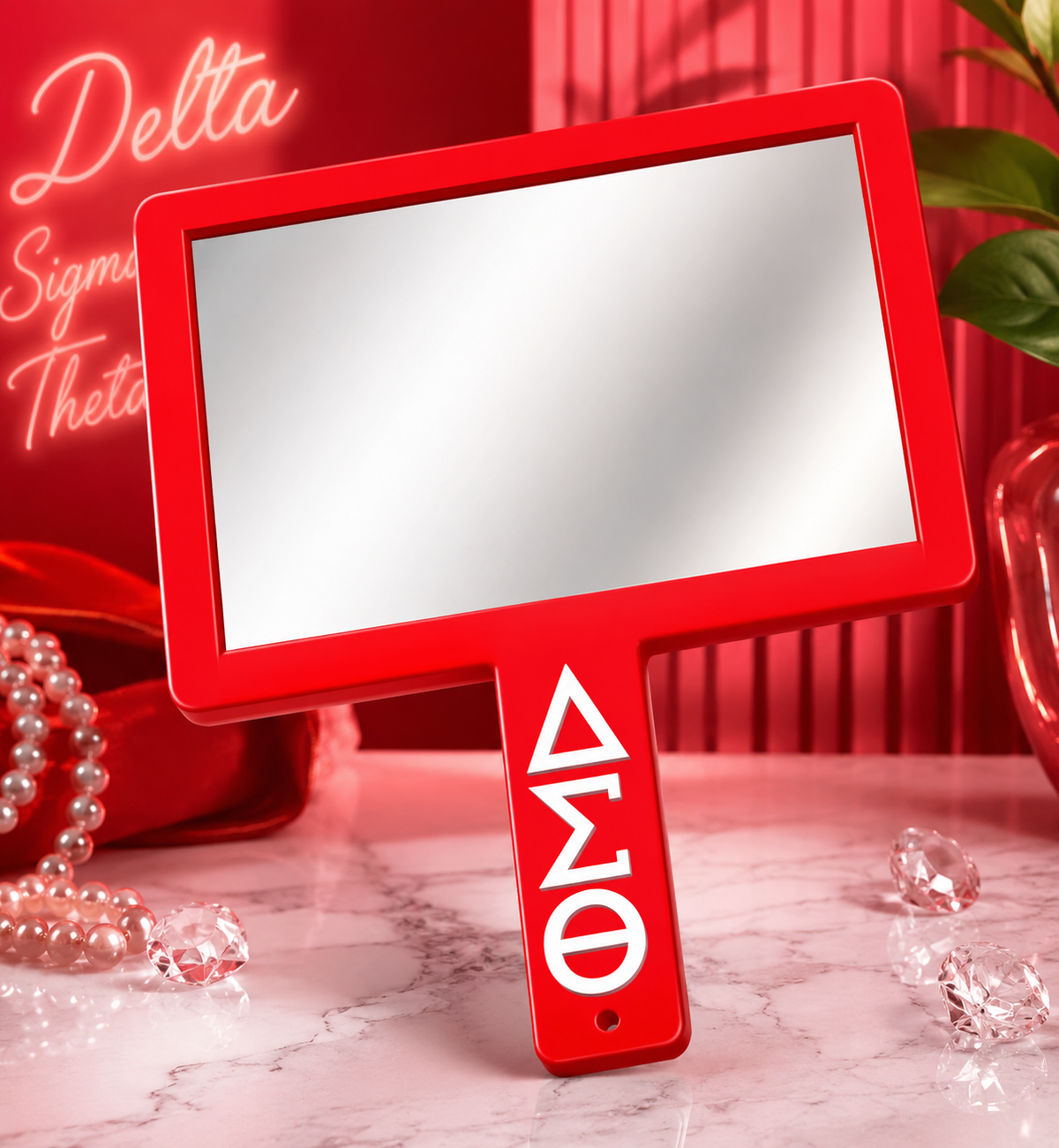 A red handheld mirror with a handle printed with the Greek letters Delta Sigma Theta in white, set on a marble surface with red and pink decorative props.