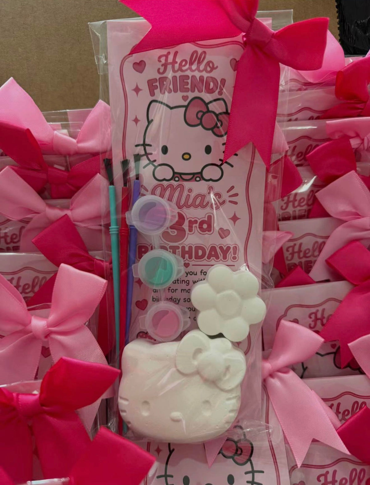 A packaged Hello Kitty-themed craft or painting kit with a pink Hello Kitty card, paintbrushes, small paint pots, and a white flower-shaped item, surrounded by pink bows and similar packages in the background.