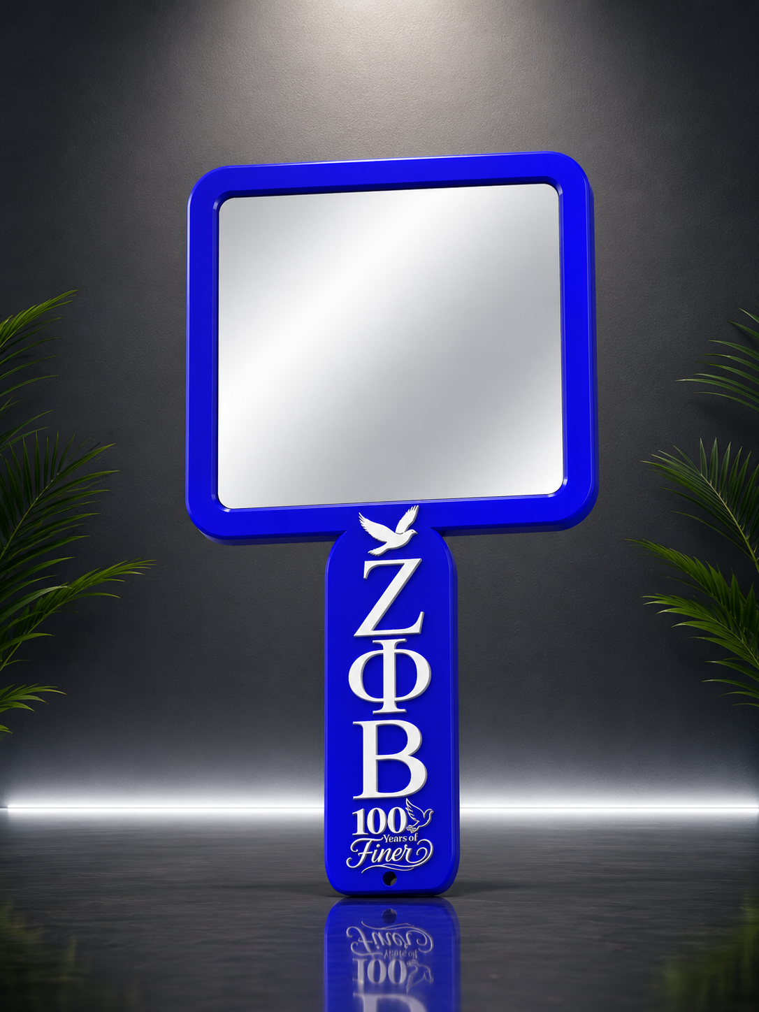 A handheld mirror with a bright blue frame and handle, featuring the Greek letters Zeta Phi Beta and '100 Finer' in white. The mirror is shown against a dark gray background with decorative green leaves on the sides.