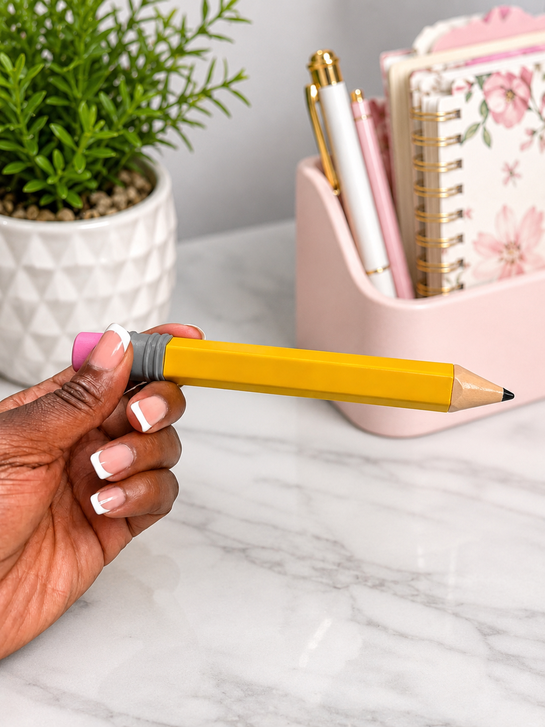 A hand with French-tip nails holds a yellow pencil with a pink eraser against a marble desk background with a pink pen cup and a potted plant.