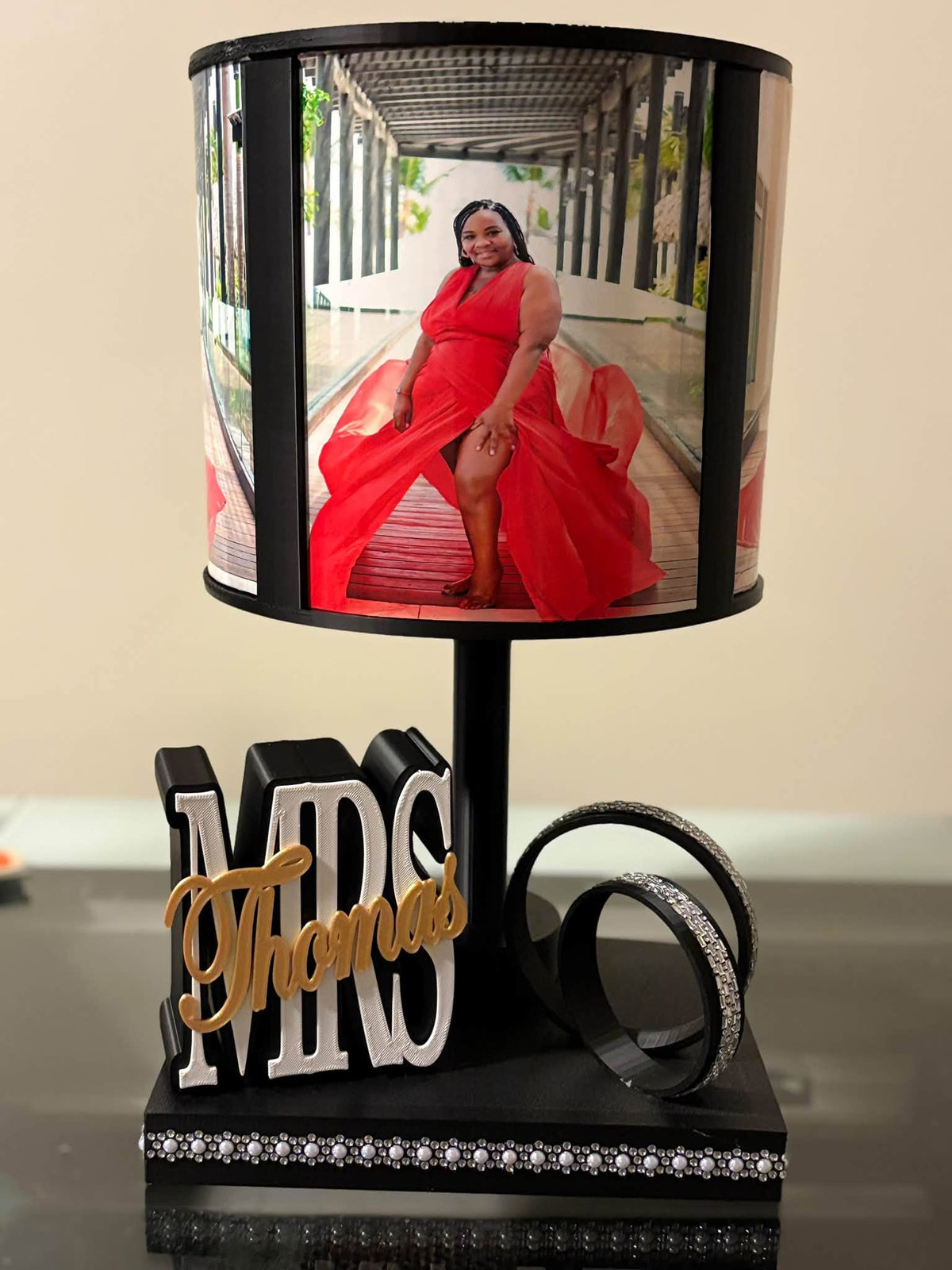 A black table lamp with a custom photo cylinder shade showing a woman in a red dress, a black base with 3D letters reading 'MRS. Thomas' in gold and white, and two black rings with silver rhinestone trim.